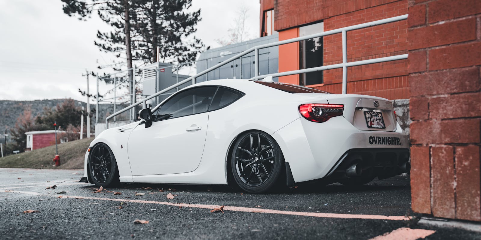 A white sports car parked next to a brick wall and metal railing, with a tree and hills in the background.