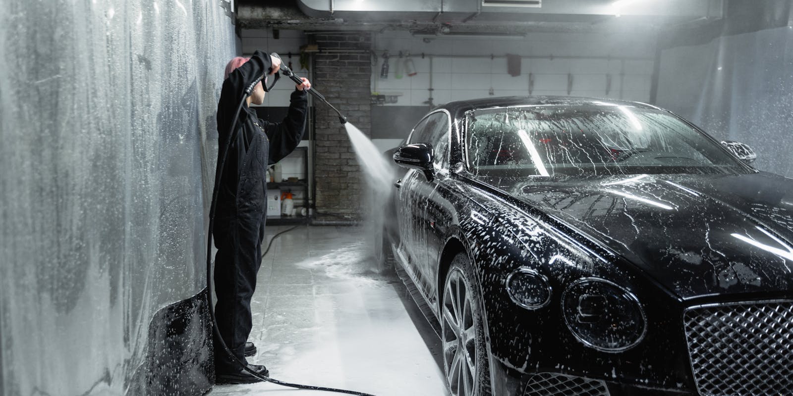 A person in black clothing power-washes a black car in a garage, creating soap suds on the vehicle.