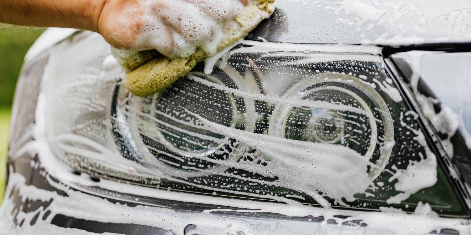 A hand cleans a car headlight with a soapy sponge.