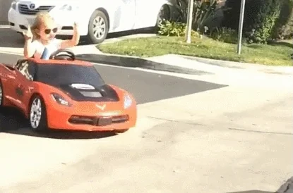 Enfant conduisant une petite voiture rouge avec des lunettes de soleil, faisant un signe rock'n'roll