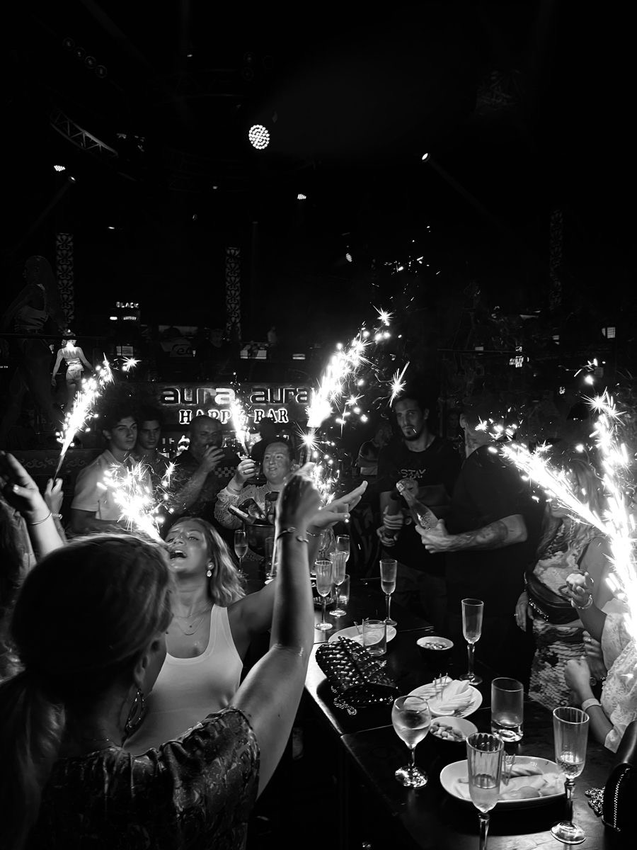 Ambiance festive dans un bar ou club avec des personnes célébrant autour de tables, des cierges magiques allumés et des verres posés, capturés en noir et blanc