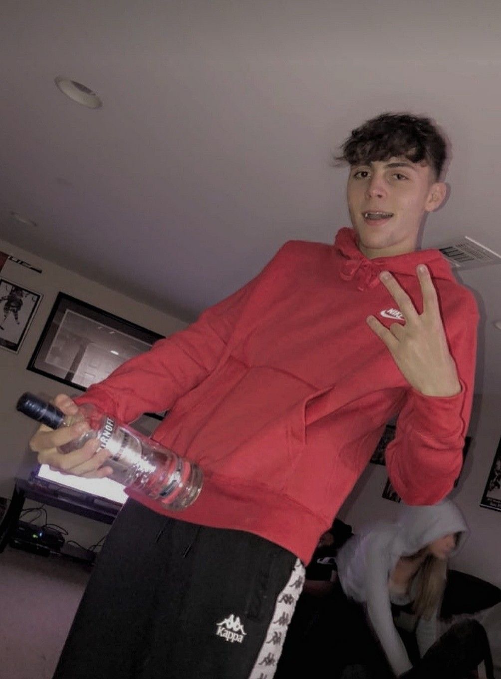 Personne portant un sweat rouge, tenant une bouteille à la main et faisant un geste de la main, dans une ambiance décontractée et festive.