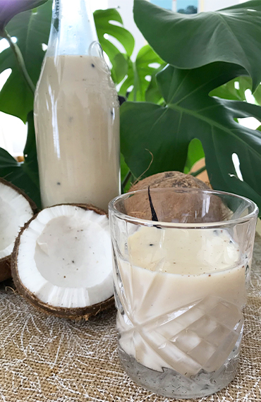 Un verre de punch coco crémeux posé devant une bouteille en verre et des moitiés de noix de coco, avec des feuilles tropicales en arrière-plan