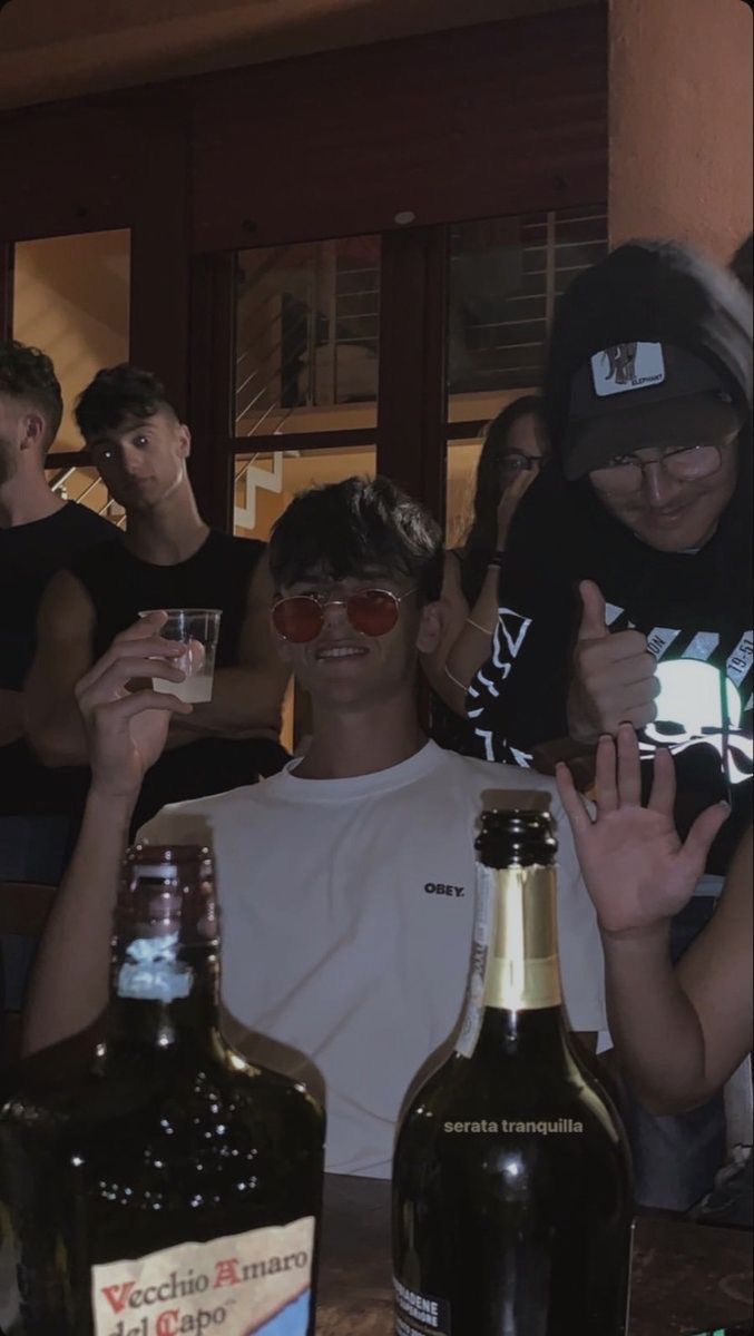 Un jeune homme portant des lunettes de soleil rouges, assis avec un verre à la main, entouré d'amis dans une ambiance festive, avec des bouteilles visibles au premier plan